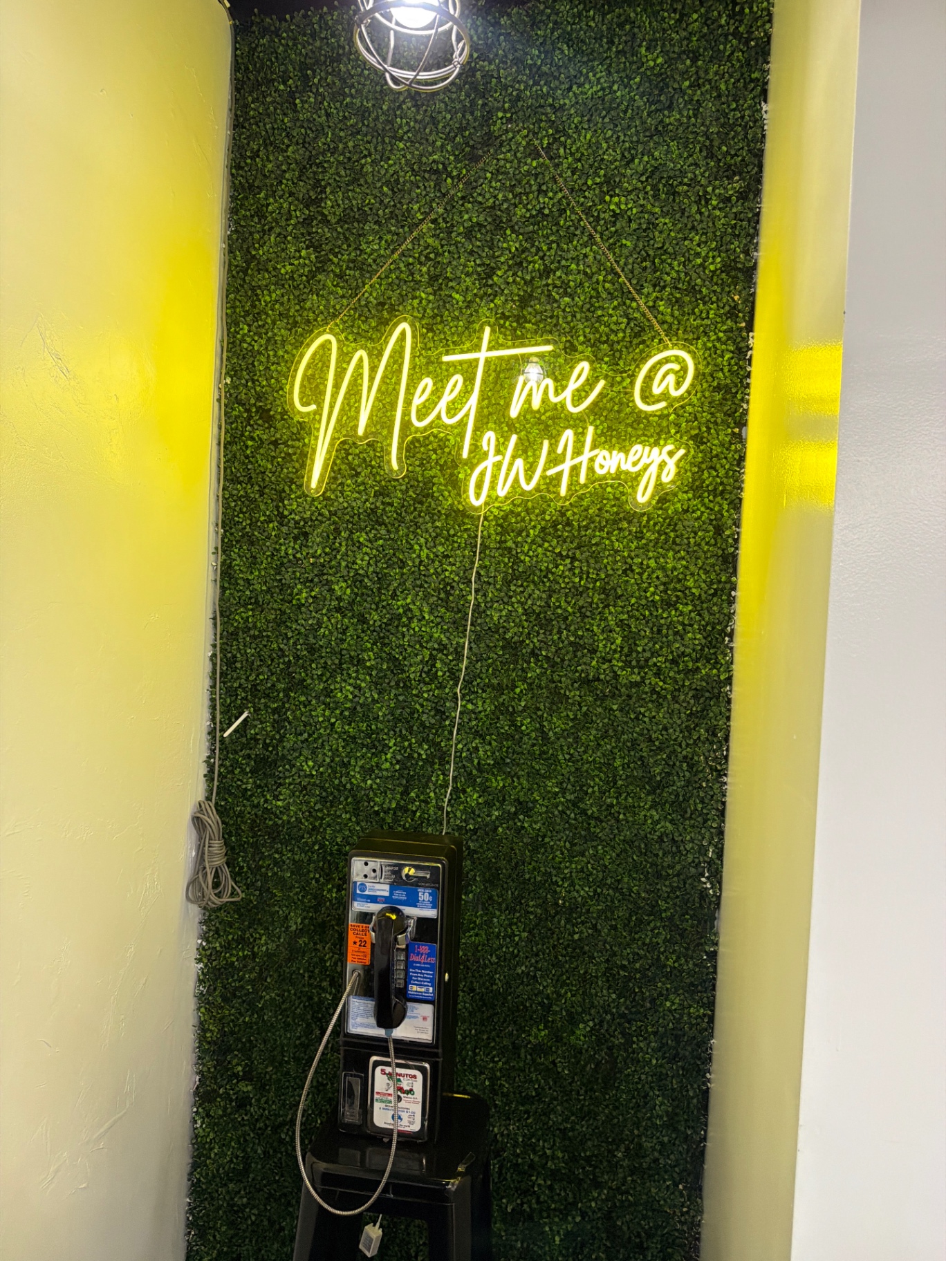 Neon sign reading Meet me @ JW Honeys on a greenery wall